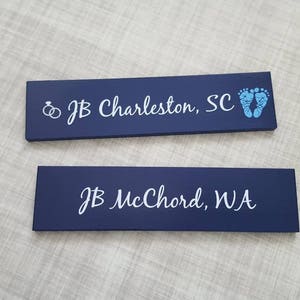 May include: Two dark blue wooden signs with white text. The top sign has a wedding ring symbol and the text "JB Charleston, SC". The bottom sign has a baby footprint symbol and the text "JB McChord, WA".