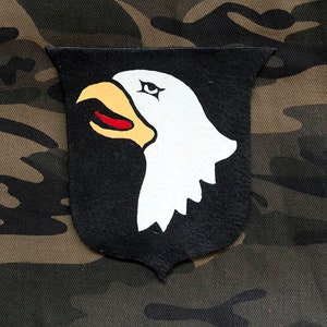 Op de afbeelding: Een zwarte leren patch met een wit adelaarhoofd ontwerp. De snavel en het oog van de adelaar zijn zwart, en het adelaarhoofd is naar links gericht. De patch is op een camouflage achtergrond.