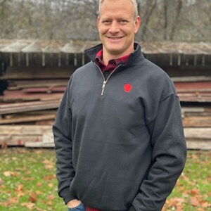 Op de afbeelding: Een man draagt een zwart mock neck sweatshirt met een rood silhouet van de staat Wisconsin. Hij draagt een blauwe jeans en een rood-zwart geruit overhemd eronder.