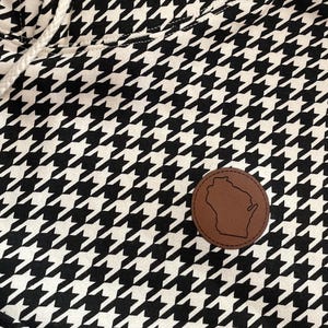 May include: Close-up of a black and white houndstooth pattern fabric. A brown leather patch with a Wisconsin state outline is sewn onto the fabric. The fabric has a drawstring and a white cord.
