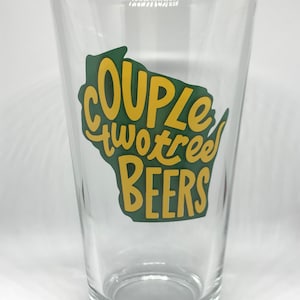 Może przedstawiać: Przezroczysty szklany kufel do piwa z zielonym i żółtym nadrukiem, na którym widnieje napis "Couple Two Trees Beers".