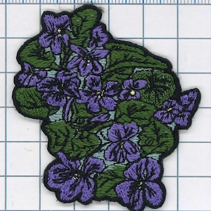 May include: Embroidered patch featuring a cluster of purple and green violets. The patch is made of fabric and has a detailed design.