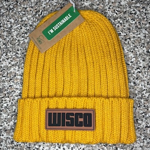 May include: A mustard yellow knitted beanie with a brown leather patch that says "WISCO" on it. The beanie has a tag that says "I'm Sustainable".