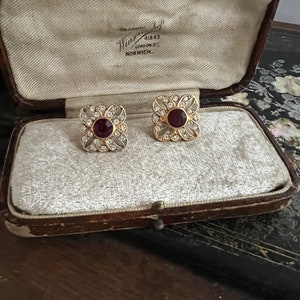 May include: A pair of gold earrings with a red stone centre and a white stone border. The earrings are in a velvet lined box with the text "Goldsmith 41843 London St. Norwich."