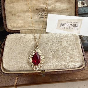 May include: A gold necklace with a teardrop pendant, set with a large red gemstone and surrounded by clear crystals. The necklace is presented in a vintage jewellery box. A tag reads "MADE WITH SWAROVSKI ELEMENTS."