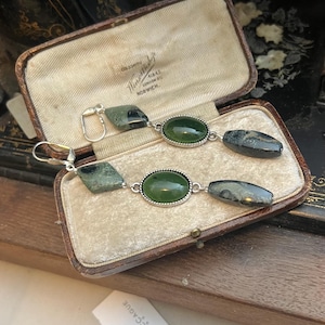 Può includere: Un paio di orecchini pendenti con pietre preziose verdi e grigie in un portagioie vintage aperto. Gli orecchini presentano pietre ovali e geometriche incastonate in metallo argentato. La scatola è foderata in tessuto color crema.