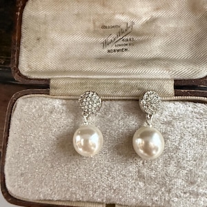 May include: A pair of drop earrings featuring round, white, lustrous faux pearls suspended from circular studs encrusted with small, clear, sparkling crystals. The earrings are displayed inside a vintage, rectangular, brown leather jewellery box.