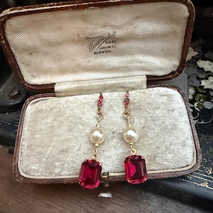 May include: A pair of drop earrings featuring a gold-tone metal setting, a small red crystal accent at the top, a white pearl bead in the center, and a large rectangular red crystal at the bottom, displayed inside a vintage brown velvet-lined box.