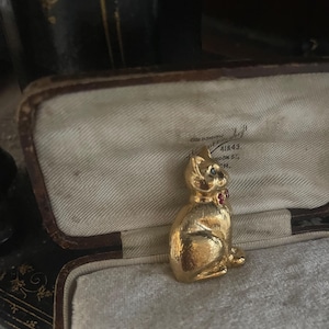 May include: A gold-coloured cat figurine with a red gem collar and blue gem eyes. The cat is sitting upright inside a vintage jewellery box with a cream-coloured interior. The box is open, revealing the cat figurine.