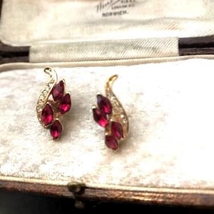 May include: A pair of vintage gold earrings with a leaf design. Each earring has several marquise-cut, deep red gemstones and small clear crystals. The earrings are displayed inside a vintage jewellery box.