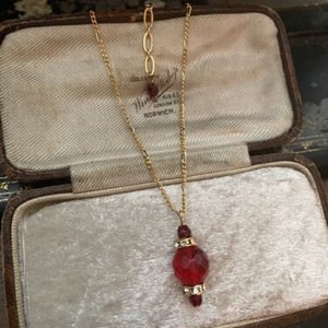 May include: A gold chain necklace with a red glass pendant. The pendant is faceted and has a small gold accent at the top. The necklace is displayed in a velvet-lined jewellery box.