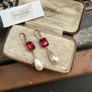 Op de afbeelding: Een paar vintage oorbellen met rode rechthoekige kristallen en peervormige parels. De goudkleurige oorbellen worden gepresenteerd in een bruine sieradendoos met een crèmekleurig interieur. Een label leest "Made with Swarovski Elements."