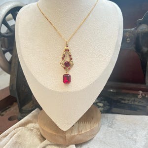 Puede incluir: Un collar de oro con un colgante de piedra roja. El colgante tiene forma de lágrima con una piedra roja más pequeña en la parte superior. El collar está en un expositor blanco.
