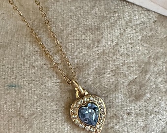 Pode incluir: Um colar de tom dourado com um pingente em forma de coração, apresentando uma pedra azul-clara central rodeada por um halo de pequenos cristais transparentes. O pingente está preso a uma corrente fina dourada, com uma etiqueta parcial escrita ELEMENTS.