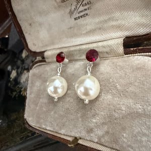 May include: A pair of drop earrings featuring a round, deep red faceted stone at the top, connected by a silver-toned metal link to a large, spherical white faux pearl bead at the bottom. The earrings are displayed inside a vintage jewellery box.