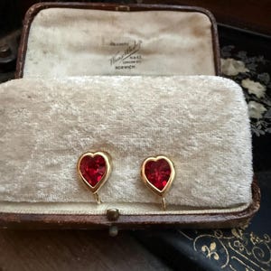 Pode incluir: Um par de brincos em forma de coração com moldura dourada e centro vermelho vibrante, exibidos numa caixa de joias vintage aberta com forro de veludo creme. A caixa é castanha com um fecho dourado.