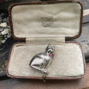 以下が含まれることがあります： 赤い宝石がアクセントの銀色の猫のブローチ。ヴィンテージのジュエリーボックスに入っています。ブローチは座っている猫のデザインです。ボックスはクリーム色の生地で裏打ちされ、内側に「Goldsmiths, Norwich」の文字があります。