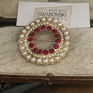 Op de afbeelding: Een goudkleurige broche met een cirkelvormig ontwerp. De broche heeft een ring van imitatieparels, rondom een ring van rode edelstenen en heldere kristallen. De speld is gemarkeerd met "Made with Swarovski Elements". De broche wordt getoond in een vintage sieradendoosje.