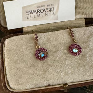 May include: A pair of gold-tone drop earrings featuring a central iridescent crystal surrounded by small pink stones. The earrings are displayed with a white card that reads MADE WITH SWAROWSKI ELEMENTS in black text, resting in a velvet-lined case.