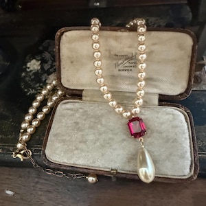 Op de afbeelding: Een ketting met een streng crèmekleurige parels, een rechthoekige donkerroze edelsteen en een enkele druppelvormige parelhanger. De ketting is voorzien van een goudkleurige metalen ketting met een karabijnsluiting en een verstelbare verlengketting.