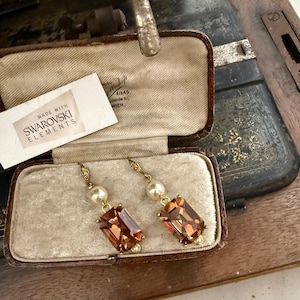 Puede incluir: Pendientes dorados con grandes cristales rectangulares color ámbar y perlas, presentados en un joyero vintage. La etiqueta dice "Made with Swarovski Elements". El joyero es marrón con interior crema.