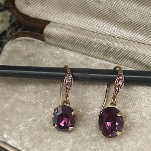 Puede incluir: Un par de pendientes colgantes dorados con gemas ovaladas de color púrpura intenso. Los pendientes tienen un pequeño grupo de gemas rosas en la parte superior del gancho. Los pendientes se muestran dentro de un joyero vintage.