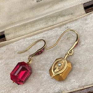 Puede incluir: Un par de pendientes de oro con piedras de vidrio rojo y un diseño de hoja de oro. Los pendientes están sobre un fondo blanco.