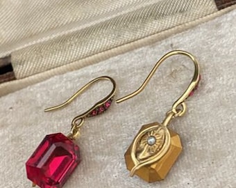 May include: A pair of gold earrings with red glass stones and a gold leaf design. The earrings are on a white background.