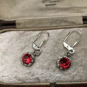 May include: A pair of silver earrings with red gemstone accents. The earrings have a simple design with a small round red stone set in a silver bezel.