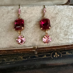 May include: A pair of drop earrings featuring a rectangular deep red crystal stone suspended above a round light pink crystal stone. The earrings have a gold-toned metal setting with a small decorative hook at the top adorned with tiny pink crystals.