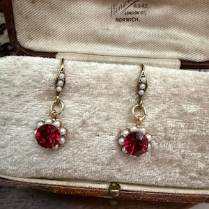 May include: A pair of antique gold-toned earrings with a red gemstone center surrounded by small pearls. The earrings are displayed in a vintage jewelry box. The box has text that reads "Goldsmith, Winson Bishop, 41&43, London St, Norwich."