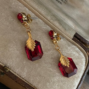 Könnte beinhalten: Ein Paar goldene Ohrringe mit roten Edelstein-Tropfen. Jeder Ohrring hat einen kleinen roten Edelstein oben und einen größeren, facettierten roten Edelstein unten. Der größere Edelstein ist von einem goldenen Blattdesign umgeben.