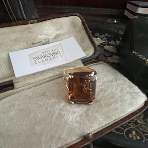 May include: A gold ring featuring a large, rectangular, amber-coloured crystal. The ring is presented in a vintage jewellery box with a cream interior. A tag reads "MADE WITH SWAROVSKI ELEMENTS".
