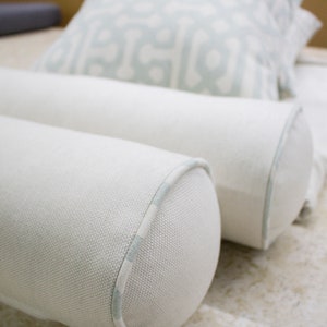 May include: Two white cylindrical pillows with a light blue trim. The pillows are made of a textured fabric and are arranged side by side.