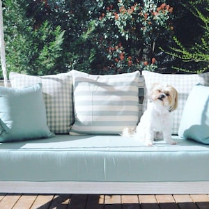 May include: A white swing bed with a light blue cushion and pillows. A small white dog with brown ears is sitting on the cushion. The swing bed is on a wooden porch with a view of green trees.