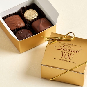 May include: A gold gift box with a gold bow and the words "Thank You" printed on the lid. The box is next to an open gold box filled with assorted chocolates.