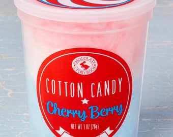 Könnte beinhalten: Ein durchsichtiger Plastikbehälter gefüllt mit rosa und blauem Zuckerwatte. Der Behälter hat einen rot-weiß gestreiften Deckel und ein rotes Etikett mit dem Text "CHOCOLATE STORYBOOK COTTON CANDY Cherry Berry NET WT 1 OZ (28g) FOR KIDS AND ADULTS".