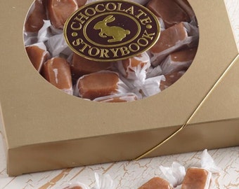 May include: A gold box with a clear window showing a variety of individually wrapped caramel candies. The box has a gold ribbon and a label that says "Chocolate Storybook" with a bunny rabbit logo.