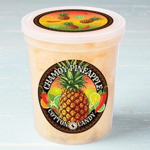 May include: A clear plastic container filled with a light yellow cotton candy. The container has a black and gold label with the text "Chamoy Pineapple Cotton Candy". The label features a pineapple, limes, chili peppers, and a floral design.