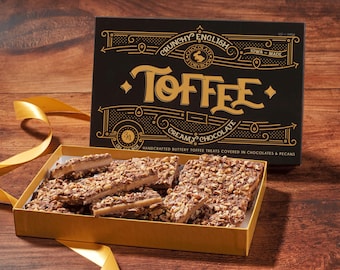 May include: A gold box filled with pieces of English toffee topped with pecans and chocolate. The box has a black lid with the words "Crunchy English Toffee" and "Creamy Chocolate" printed on it.