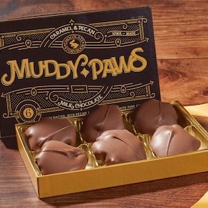 May include: A box of six milk chocolate caramel and pecan candies from Muddy Paws Chocolate Storybook. The box is gold with a black lid featuring the company logo and the text "Caramel & Pecan, Iowa-Made, Muddy Paws, Milk Chocolate, Crafted, Rich Pecan & Pure", and a number "6".