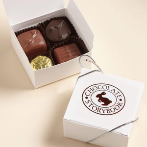 May include: Two white boxes of chocolates. One box is open, revealing four assorted chocolates. The other box is closed, with a logo that says "Chocolate Storybook" and a rabbit illustration. The chocolates are various shades of brown.