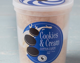 Könnte beinhalten: Ein durchsichtiger Plastikbehälter gefüllt mit Cookies & Cream Zuckerwatte. Der Behälter hat einen blauen und weißen Spiraldeckel und ein blaues Etikett mit einem weißen Milchspritzer und drei schwarzen und weißen Keksen. Das Etikett lautet "Cookies & Cream Cotton Candy" und "net wt. 1.75 oz (50g)".