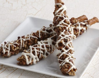 May include: Three long pretzel rods coated in chopped brown nuts and drizzled with a white icing lattice pattern. The treats are arranged in a cluster on a square white plate, showcasing the textured nut coating and the bright white glaze.