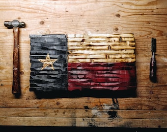May include: A wooden Texas flag with a white star on a blue background, a red stripe, and a white stripe. The flag is made of individual pieces of wood that have been cut and arranged to create a textured look. The flag is displayed on a wooden surface with a hammer and a chisel.