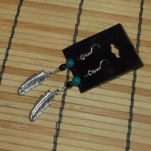 May include: Silver feather earrings with teal and black beads. The earrings have a hook closure and are displayed on a black card. The feathers are detailed with etched lines, and the beads are round.