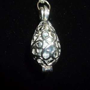 May include: A silver-colored pendant necklace with a teardrop-shaped cage design. The cage has an open filigree pattern, and it holds a light-colored, possibly translucent, stone. The necklace is on a black background.