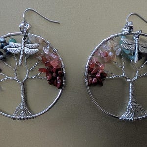 May include: A pair of silver-toned wire earrings. Each earring features a circular design with a tree of life motif, adorned with colorful gemstones and a dragonfly charm. The earrings are designed with a hook closure.