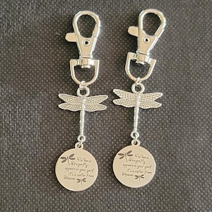 May include: Two silver-colored keychains with dragonfly charms. Each keychain has a lobster clasp, a dragonfly charm, and a circular pendant with the text "When a dragonfly appears in your yard, it's a visitor from Heaven."