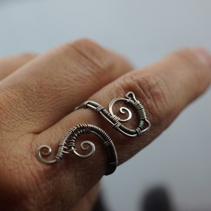 May include: A silver-colored wire ring with a unique, swirling design. The ring features a spiral and curved wire elements, creating an artistic and handcrafted look. The ring is worn on a finger.